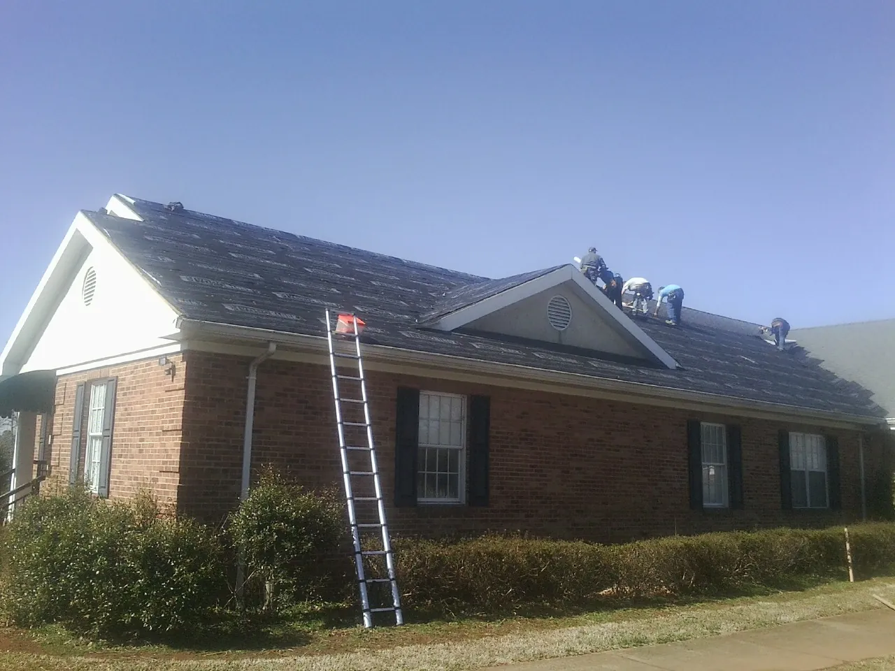 Crew tearing shingles off a brick ranch roof with a ladder set — Emergency Roof Repair work by our crew in Roggen