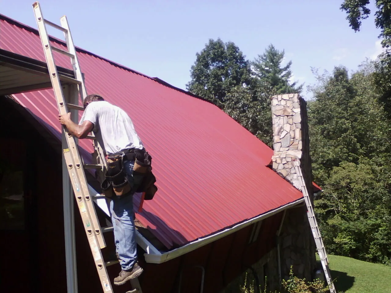 Roofer on a ladder beside a red metal roof and stone chimney — Soffit & Fascia Repair by Roofing Brothers in Roggen